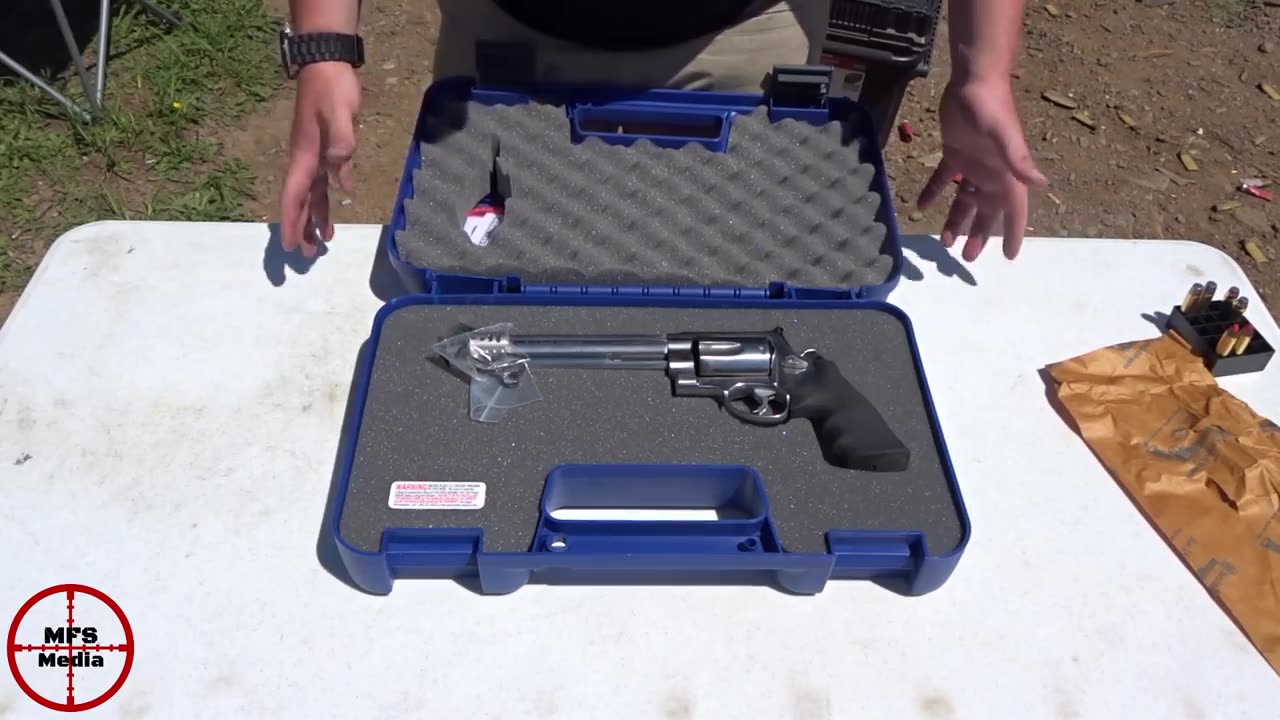 The Smith & Wesson 460XVR in its blue factory case with foam lining, the spare muzzle brake in a small bag at the muzzle, and a box of ammunition on the table
